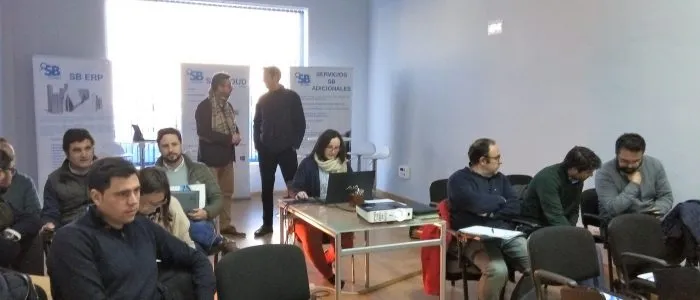 Asistentes sentados en la sala, con carteles de SB ERP y Servicios SB Adicionales al fondo y dos hombres hablando de pie