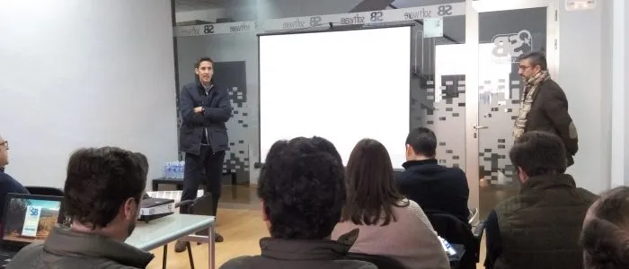 Técnicos sentados de espaldas escuchan a los dos ponentes, de pie a ambos lados de la pantalla, en la sala de SB Software