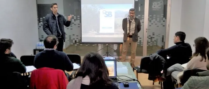 Dos ponentes de pie a ambos lados de la pantalla, que muestra el acceso a la plataforma con el logotipo de SB sobre un olivar
