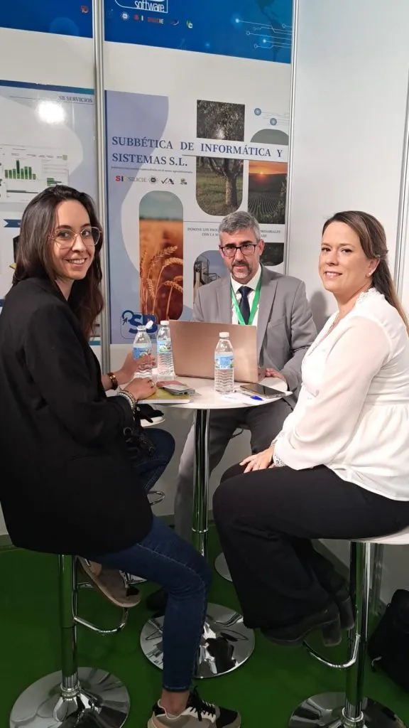 Dos mujeres y un hombre reunidos en una mesa alta del stand, con un portátil abierto y botellas de agua
