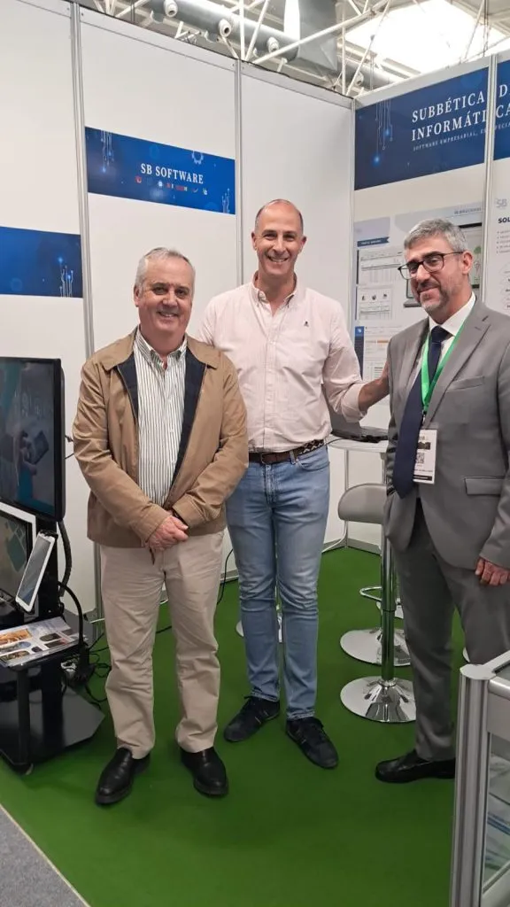 Tres hombres posan sonrientes en el stand de SB Software, junto a las pantallas y las mesas altas