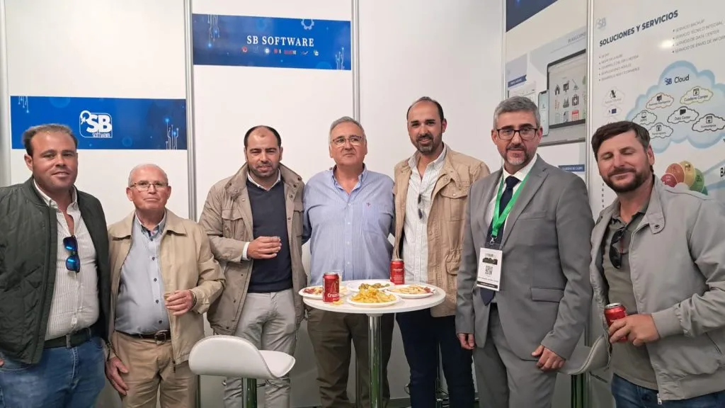 Siete hombres posan alrededor de una mesa alta con aperitivos bajo los rótulos de SB Software