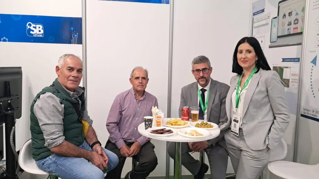 Tres hombres sentados y una mujer de pie comparten un aperitivo en una mesa alta del stand de SB Software