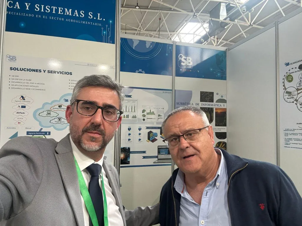 Selfi de dos hombres en el stand de SB Software, con el panel de soluciones y servicios al fondo