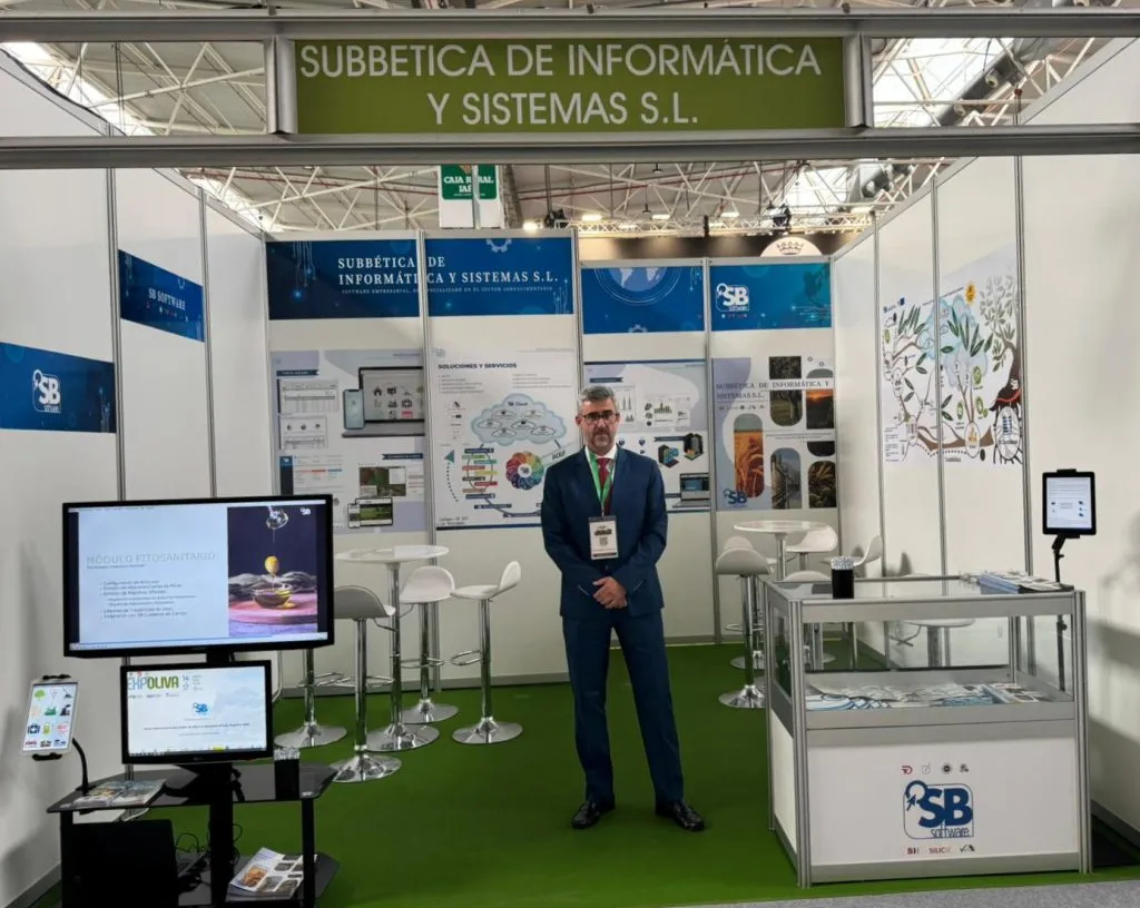 Un hombre con traje y acreditación espera en el centro del stand de SB Software, entre pantallas y mesas altas
