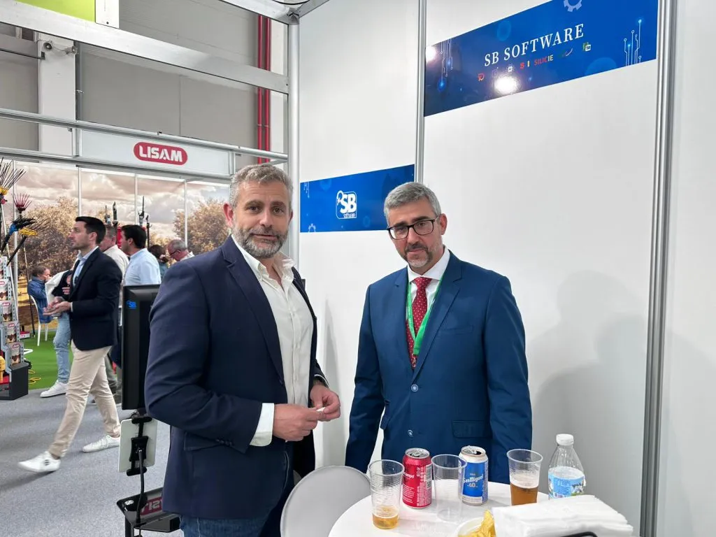 Dos hombres posan junto a una mesa alta con bebidas bajo el rótulo de SB Software, con visitantes de la feria al fondo